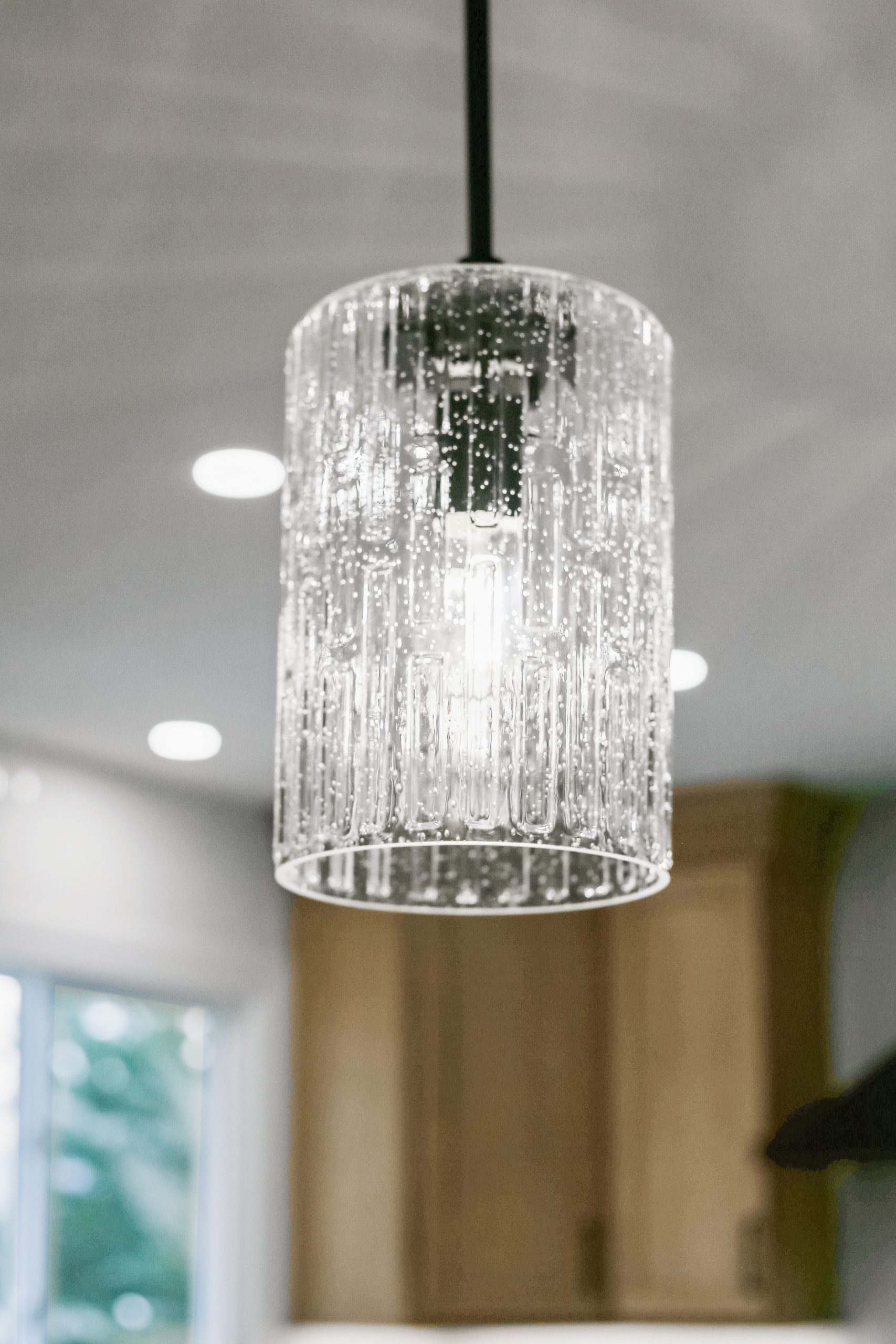 A close-up of a modern pendant light with a cylindrical, textured glass shade, illuminated and hanging from the ceiling in a bright room with blurred background elements.