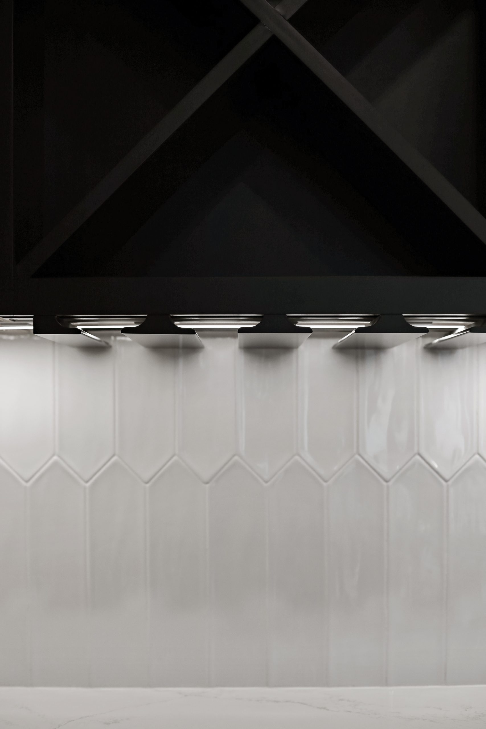 A close-up of a modern kitchen backsplash with glossy, light gray hexagonal tiles and a black cabinet above, illuminated by under-cabinet lighting.