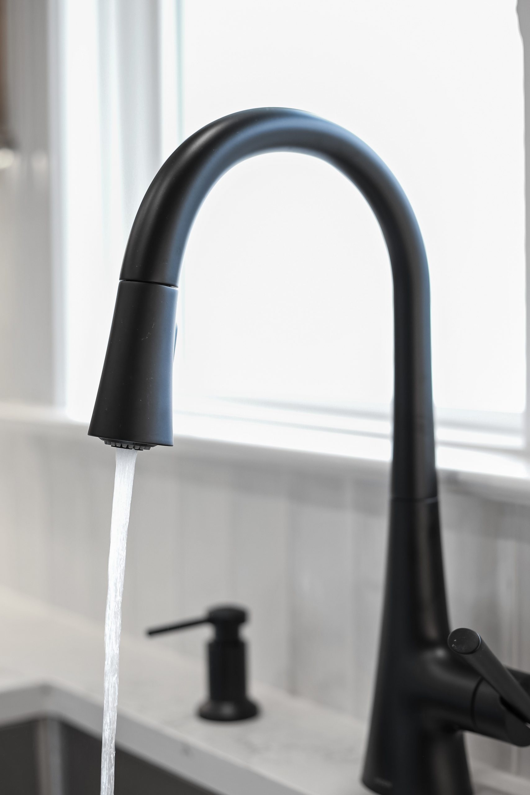 A modern black kitchen faucet with water running, set against a bright window and white tiled backsplash. A soap dispenser is visible in the background near the sink.