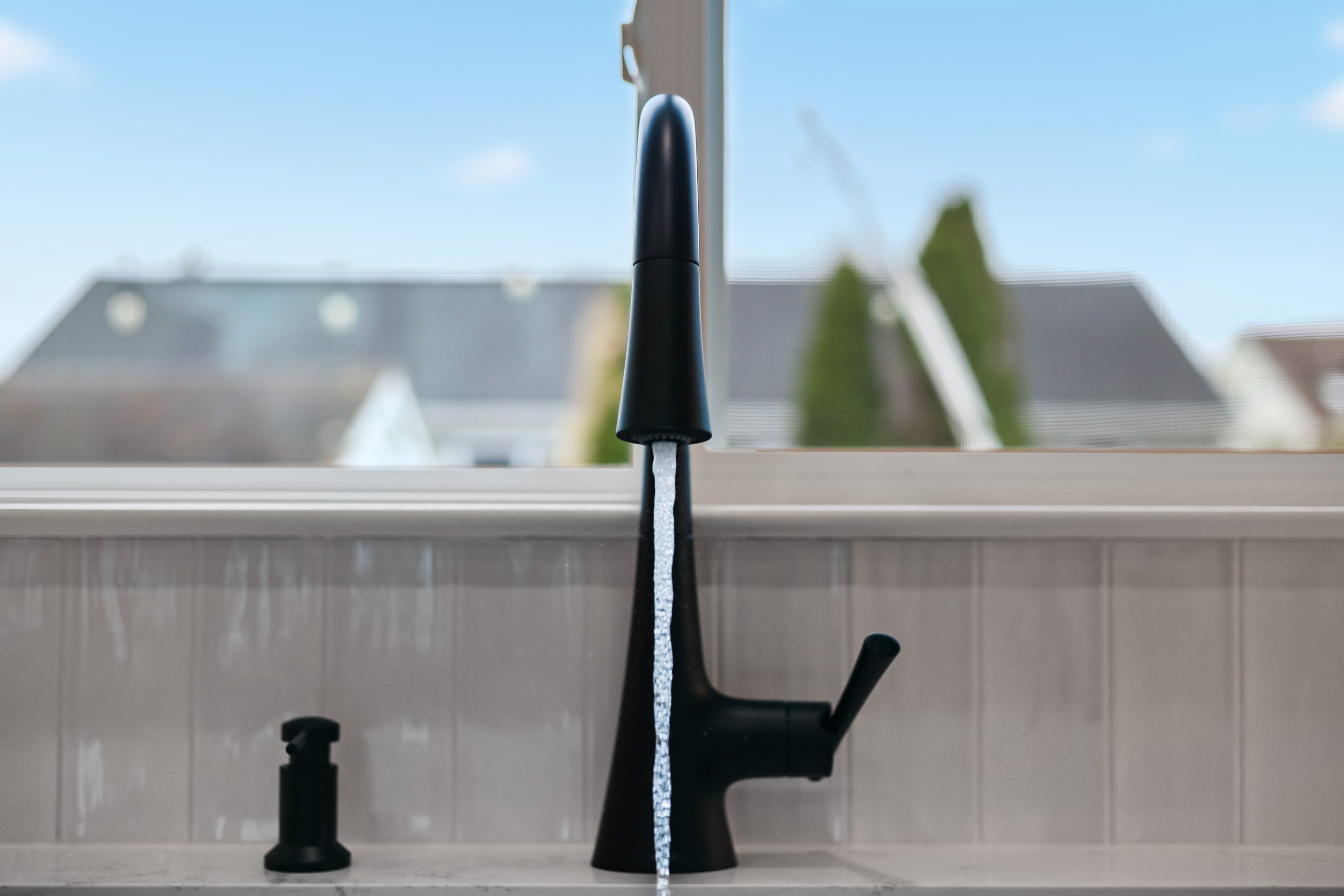 A modern black kitchen faucet with water running, set in front of a window with an outdoor view of houses and trees. A black soap dispenser is on the left side of the faucet.