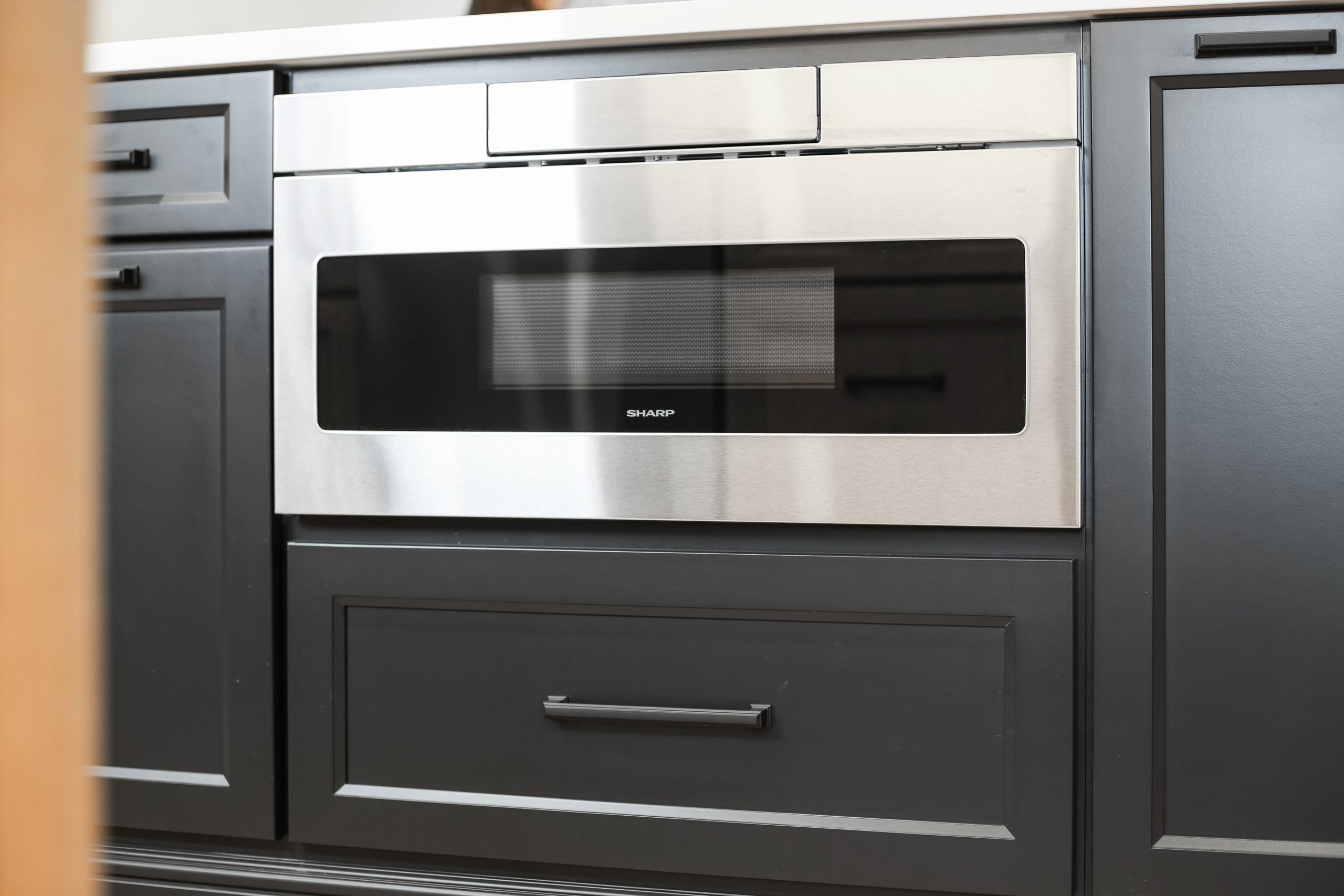 A built-in stainless steel microwave oven is installed within black kitchen cabinetry, with a drawer below and cabinets on either side.