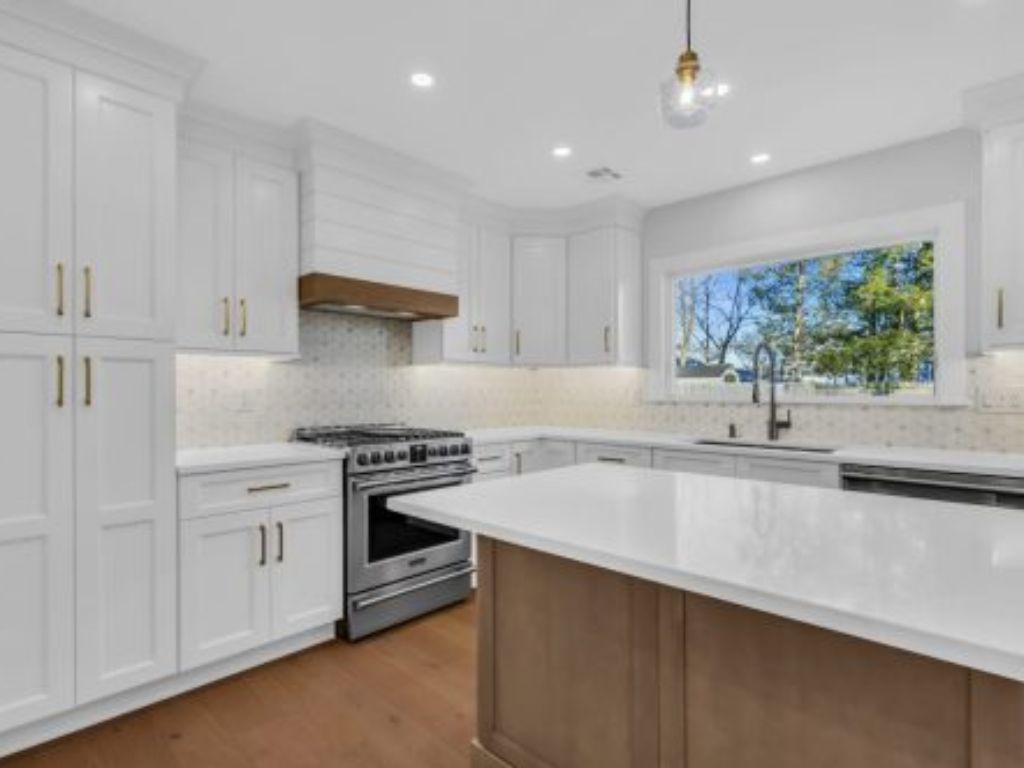 Modern kitchen remodeling with white cabinets and gold handles, stainless steel oven, large island, hardwood floor, and window view of trees.
