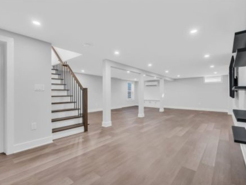 Spacious basement with light wood flooring, white walls, recessed lighting, and a staircase with dark railings. Shelves on the right wall add a modern touch.