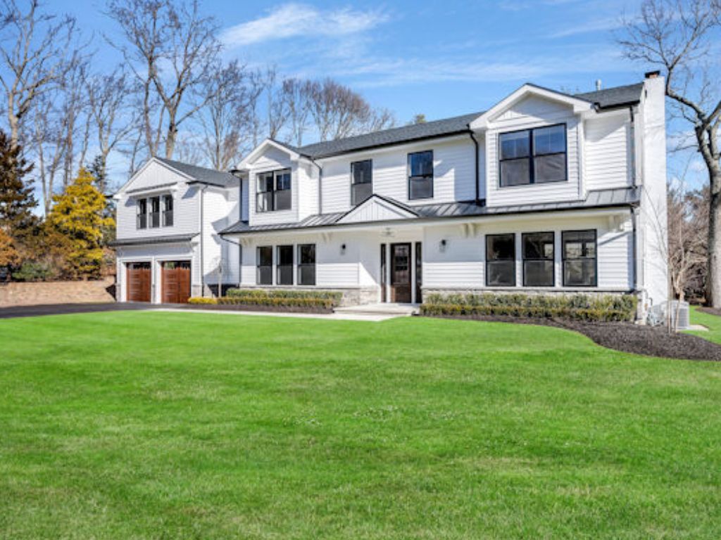 A large, modern two-story white house with black trim and multiple windows—recently transformed by a full home renovation in New Jersey—sits on a spacious, manicured green lawn under a clear blue sky. An attached garage features elegant wooden doors.
