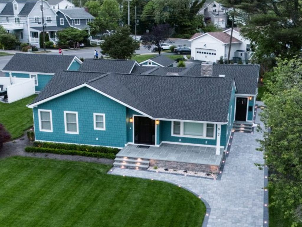 Aerial view of a modern blue house with white trim, featuring recent roof and siding upgrades, a dark shingle roof, paved driveway, manicured green lawn, and landscaped walkway. Nearby houses and trees are visible in the background.